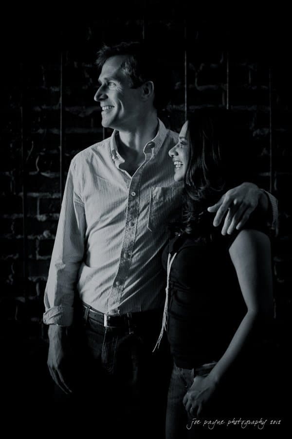 unc chapel hill engagement session ~ thelsa and phil A black-and-white photo of a man and a woman standing close and smiling. The man has his arm around the womans shoulder. They are looking slightly to the left against a brick wall background.