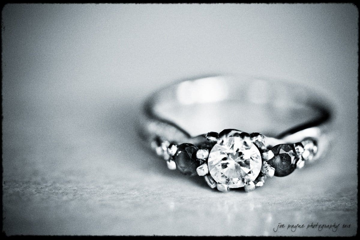 A close-up black and white photograph of an ornate ring featuring a central round diamond flanked by smaller dark stones set on a smooth band. The background is softly blurred to emphasize the rings details.