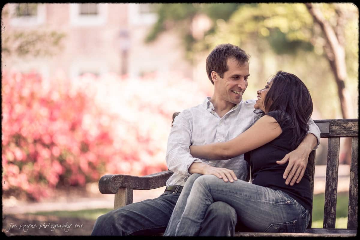 unc chapel hill engagement session ~ thelsa and phil A couple sits on a park bench, smiling and embracing each other. They are surrounded by pink flowers and greenery, creating a peaceful and romantic atmosphere.