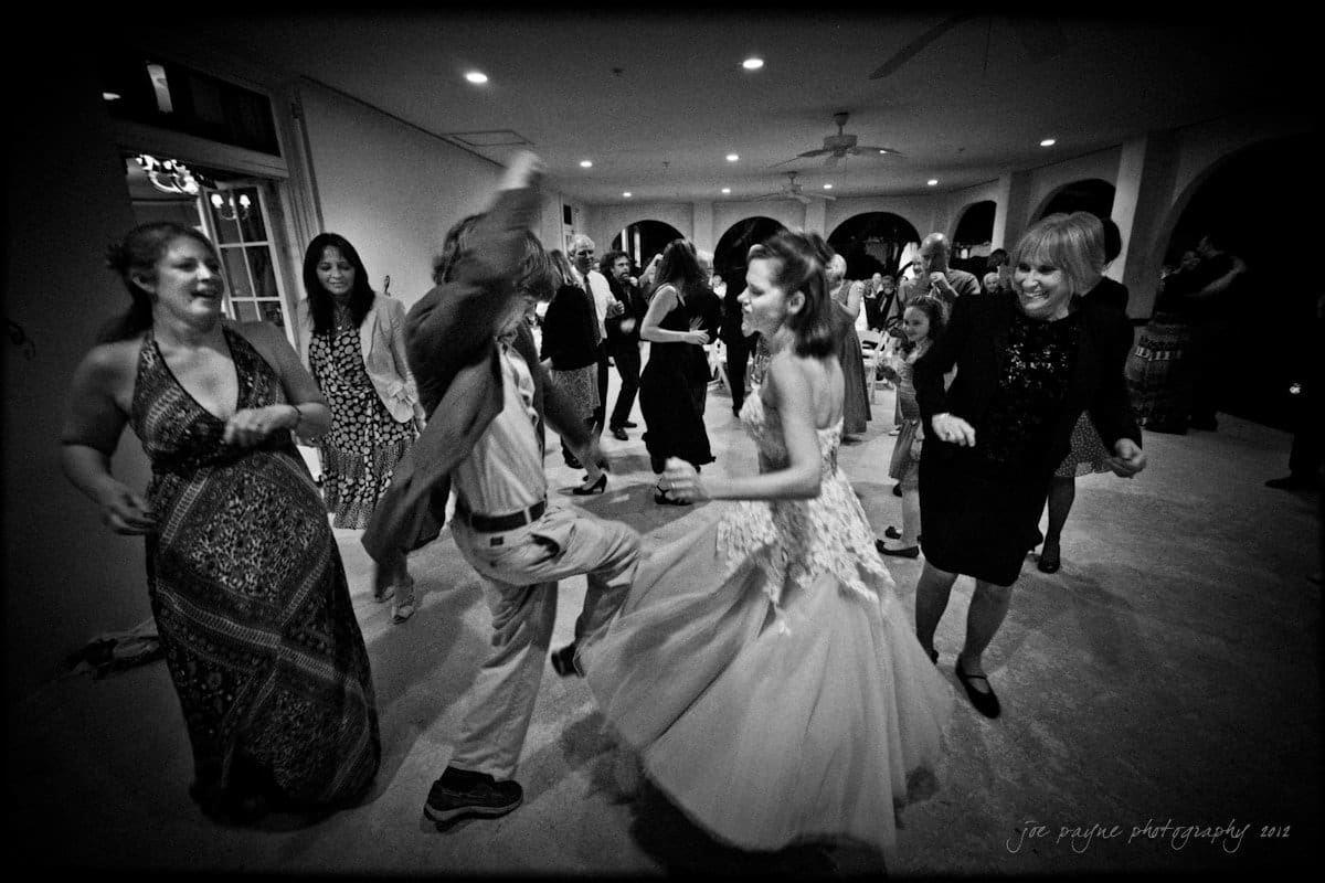 lake lure north carolina wedding photography ~ shannon and paul A lively black and white photo captures a group of people dancing energetically at a party. In the foreground, a young woman in a gown dances with a man in a hat. Smiling onlookers surround them, filling the room with joyful movement.