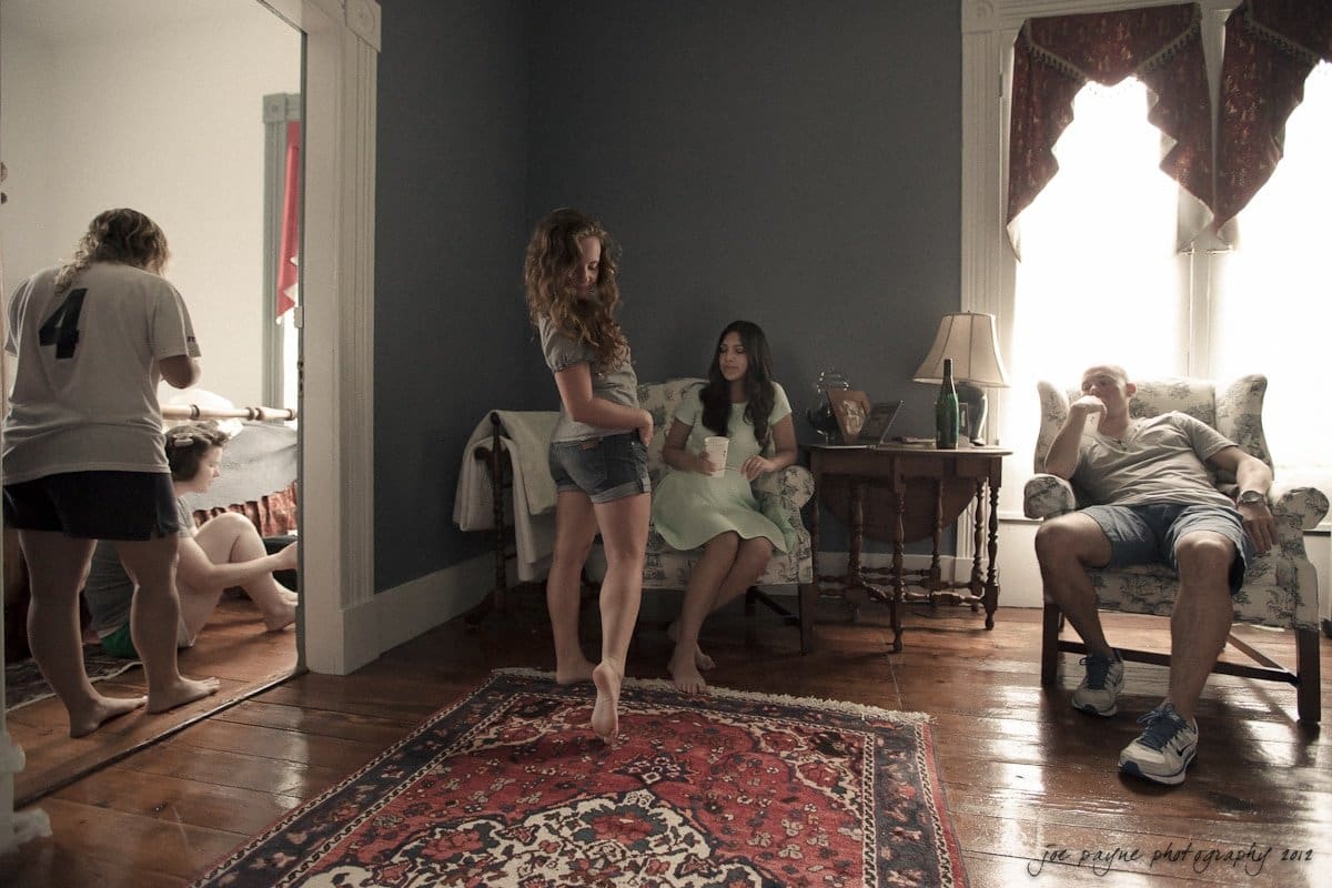 A cozy living room with a vintage rug and comfortable furniture. Four people are relaxing; one is seated on a chair, two are interacting in the center, and another is kneeling in a doorway. Warm lighting fills the space.