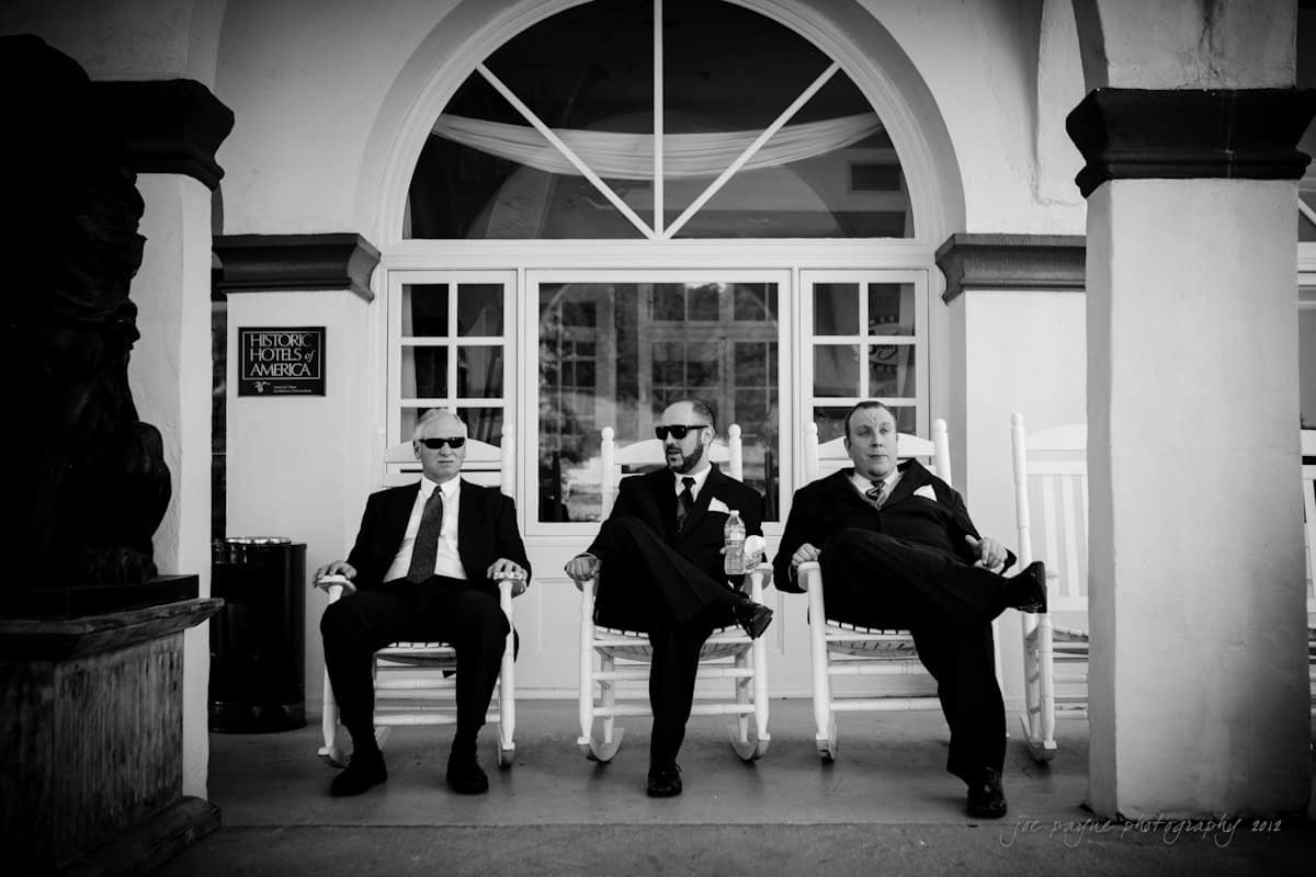 lake lure north carolina wedding photography ~ shannon and paul Three men in suits and sunglasses sit on rocking chairs outside a white building. The photo is in black and white, giving it a classic feel. They have a relaxed, confident posture.