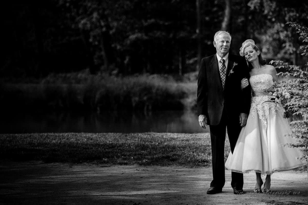 lake lure north carolina wedding photography ~ shannon and paul A black and white photo of a bride and an older man walking arm in arm on a path. The bride holds a bouquet and wears a strapless dress. The background features a pond and trees, creating a serene atmosphere.