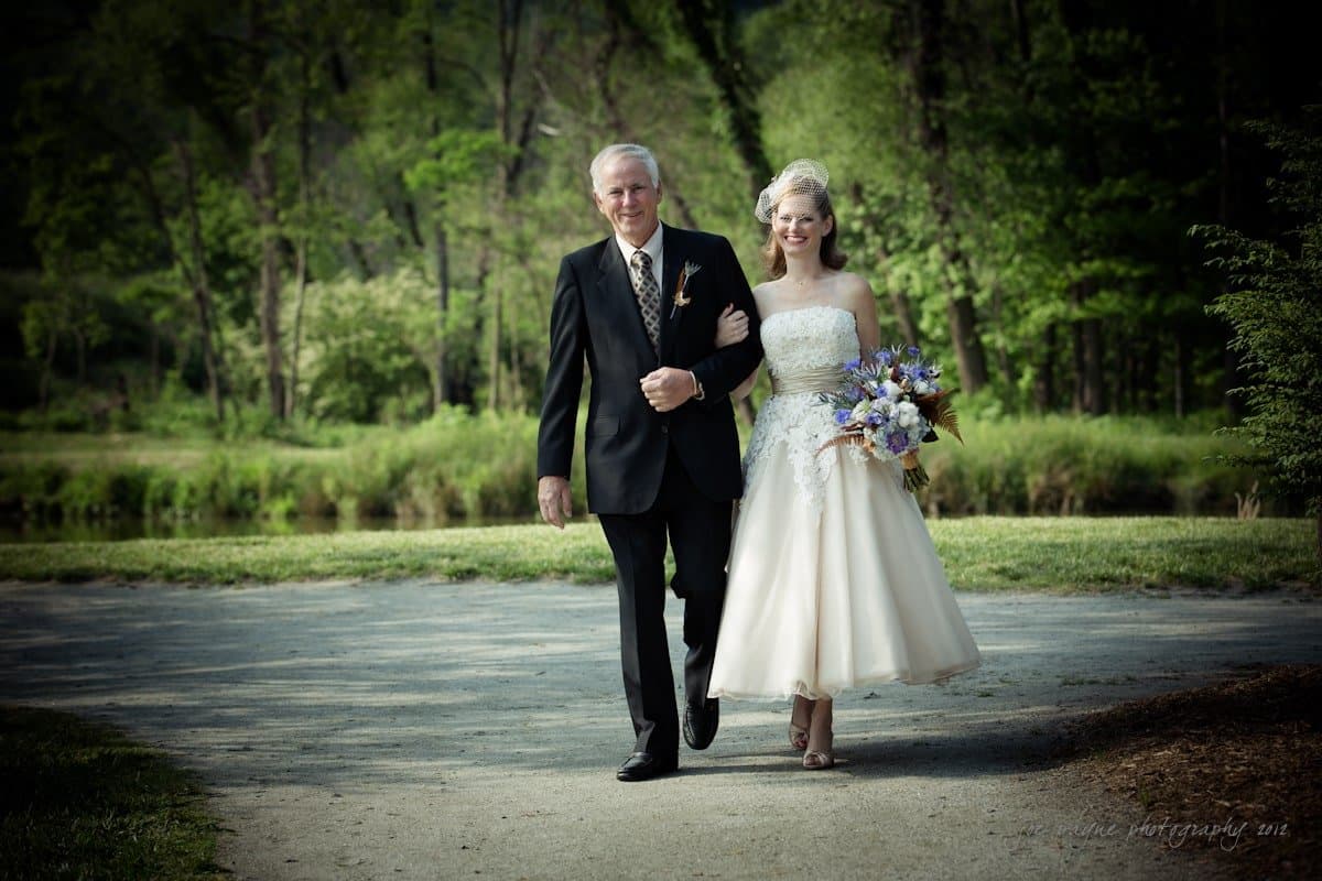 lake lure north carolina wedding photography ~ shannon and paul A bride in a white dress and veil holds a colorful bouquet as she walks arm in arm with an older man in a suit and tie. They are outdoors on a path surrounded by lush green trees and a small body of water.