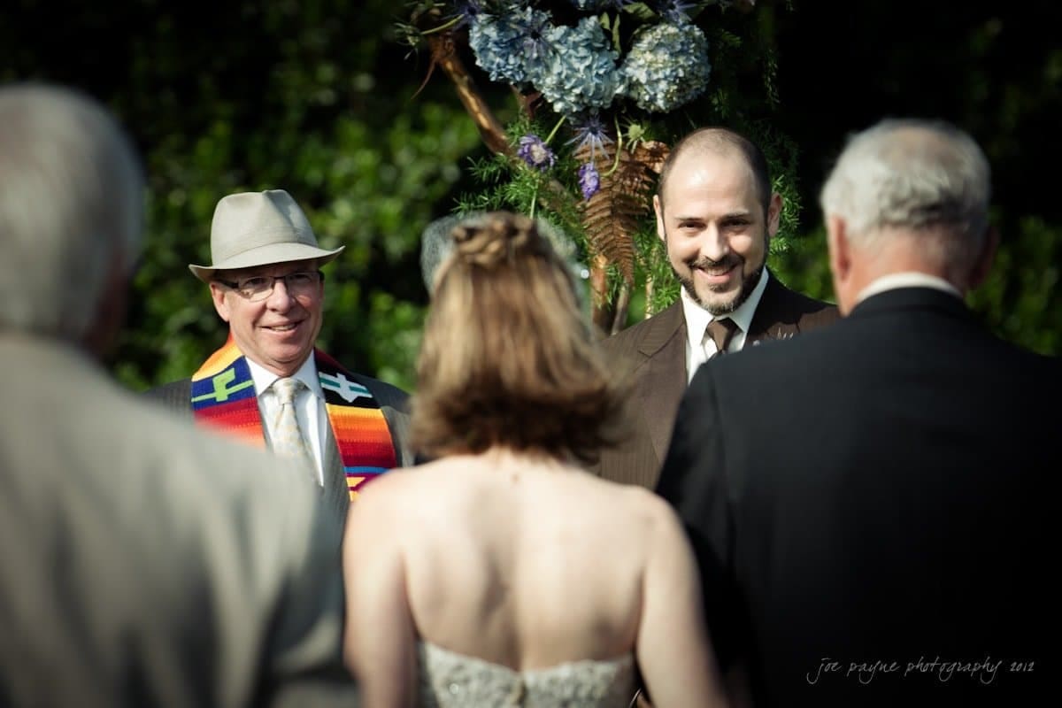 lake lure north carolina wedding photography ~ shannon and paul A bride and groom stand before an officiant in a colorful stole, smiling during an outdoor wedding ceremony. The scene is warmly lit, with guests in the foreground and floral decorations in the background.