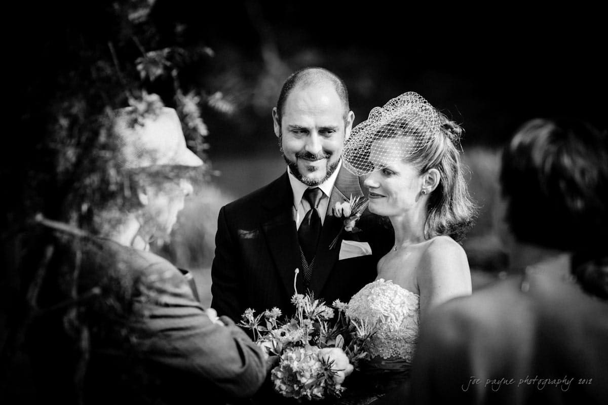A bride and groom, dressed elegantly, stand facing a celebrant during an outdoor wedding ceremony. The bride holds a bouquet and wears a birdcage veil. Lush greenery surrounds them, and another person is partially visible in the foreground.
