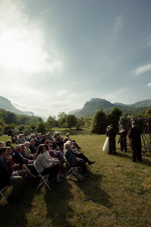 lake lure north carolina wedding photography ~ shannon and paul A wedding ceremony takes place outdoors on a sunny day, with guests seated on folding chairs. The bride and groom stand before a natural backdrop of mountains and trees. The sky is partly cloudy, and a small group of people look on attentively.
