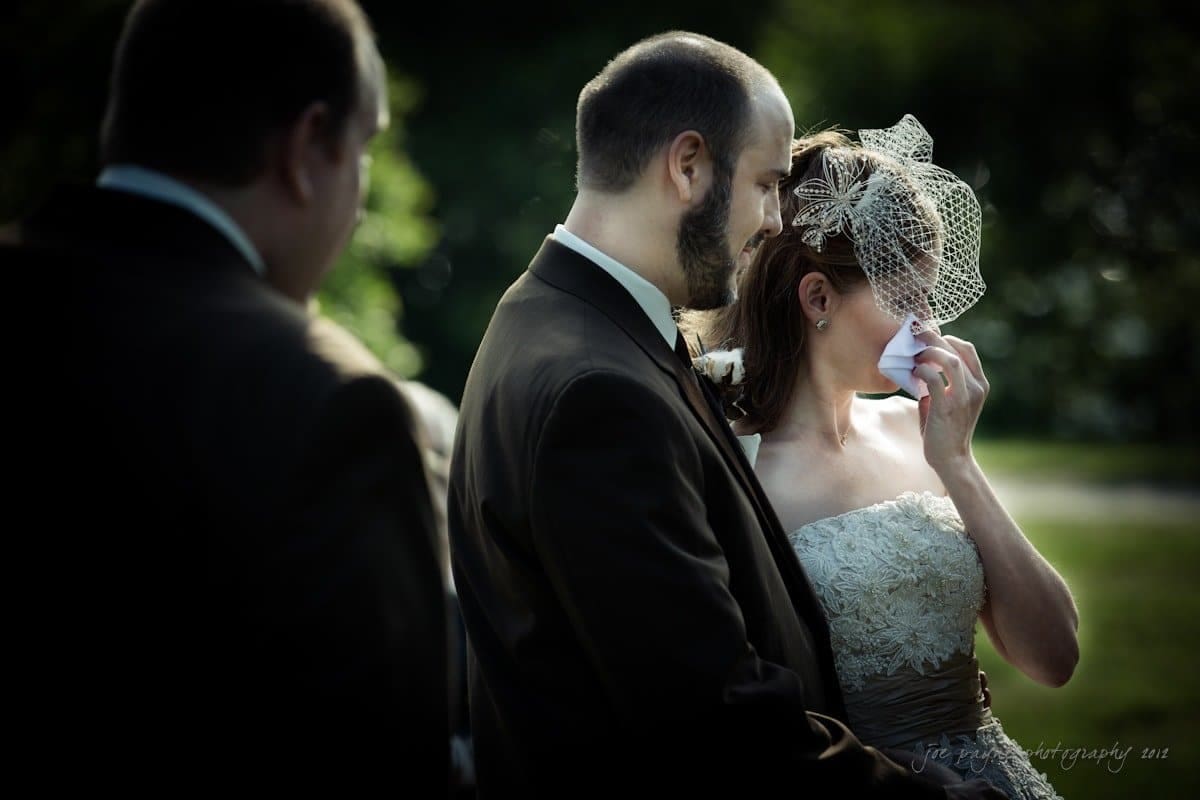 lake lure north carolina wedding photography ~ shannon and paul A bride wipes her eyes with a tissue while standing next to a groom in a dark suit. They are outdoors, with another person partially visible. The mood is emotional and intimate.