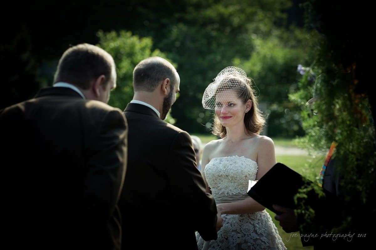 lake lure north carolina wedding photography ~ shannon and paul A bride and groom stand facing each other during a wedding ceremony outdoors. The bride, in a white dress with a netted veil, smiles softly. The officiant holds a book, while another person stands nearby. Sunlight filters through the surrounding trees.