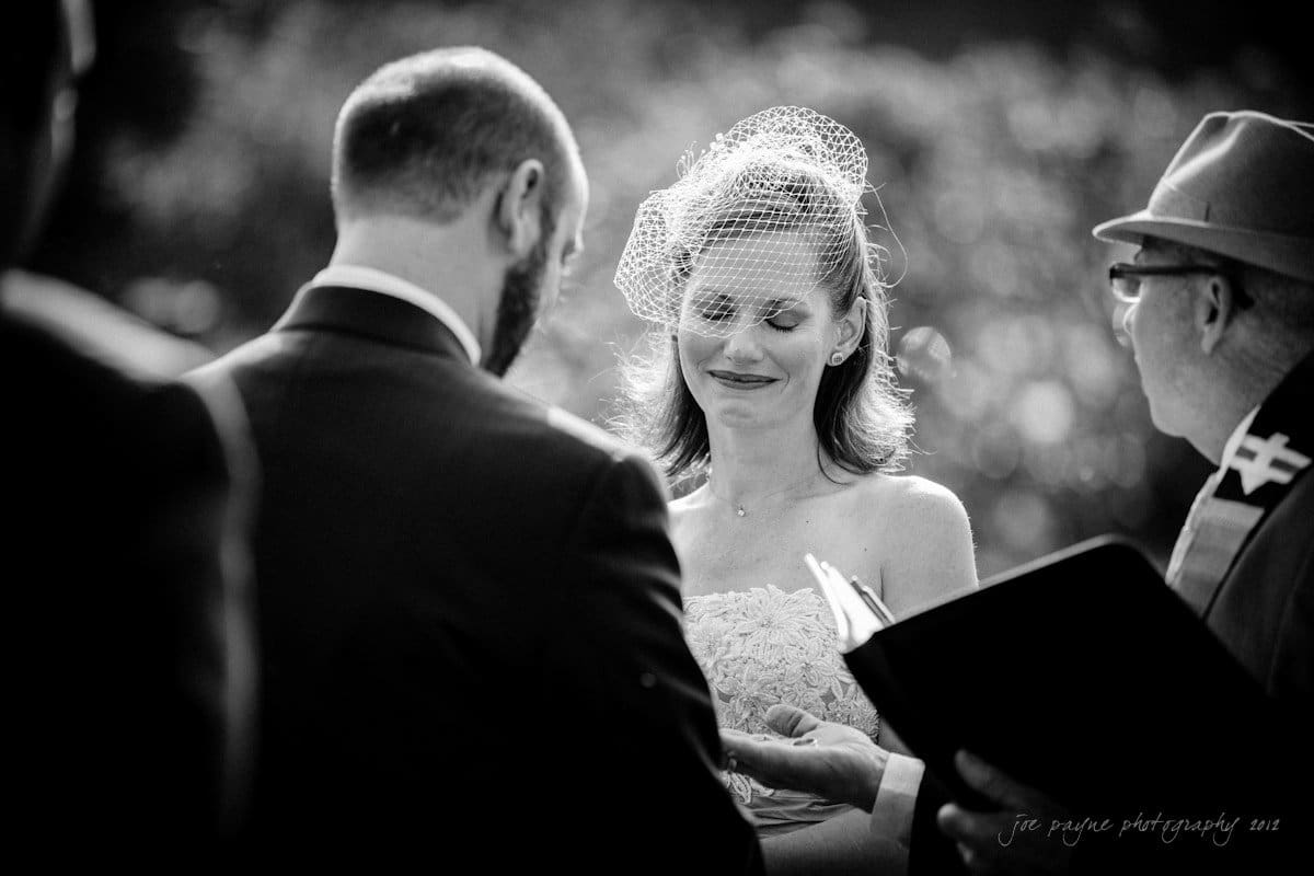 lake lure north carolina wedding photography ~ shannon and paul A black-and-white image of a wedding ceremony outdoors. The bride, wearing a strapless dress and birdcage veil, smiles at the groom. The officiant is reading from a book. The scene is illuminated by natural light.