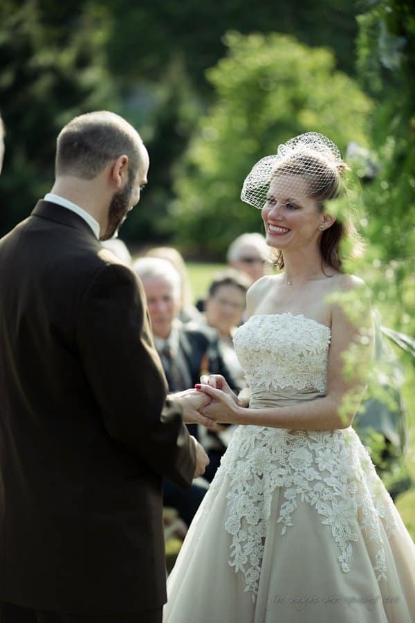 lake lure north carolina wedding photography ~ shannon and paul A couple in formal attire exchanges vows outdoors. The bride wears a lace gown with a birdcage veil, smiling at her partner who is in a dark suit. Guests are seated in the background amidst greenery.