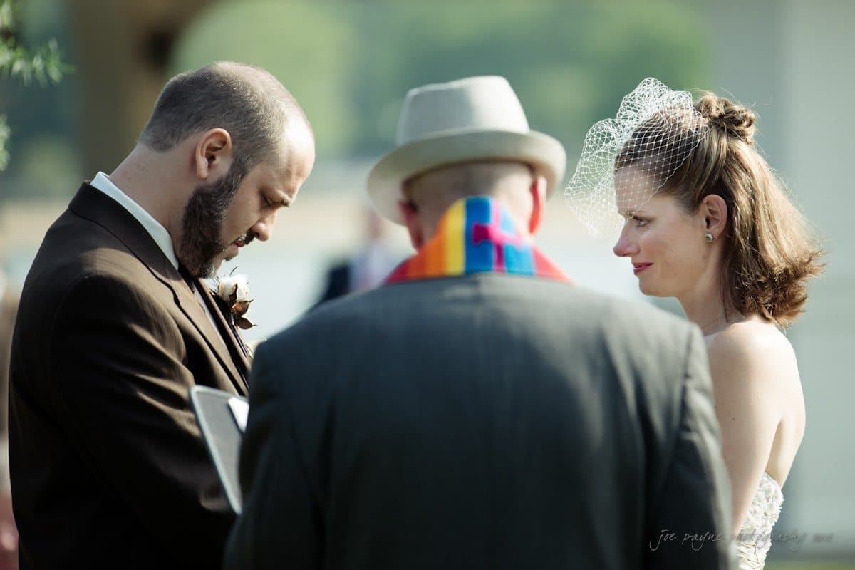 lake lure north carolina wedding photography ~ shannon and paul A couple is getting married outdoors, standing before an officiant with a colorful scarf. The groom wears a suit, and the bride wears a white dress with a birdcage veil. Trees and a blurred background suggest a natural setting.