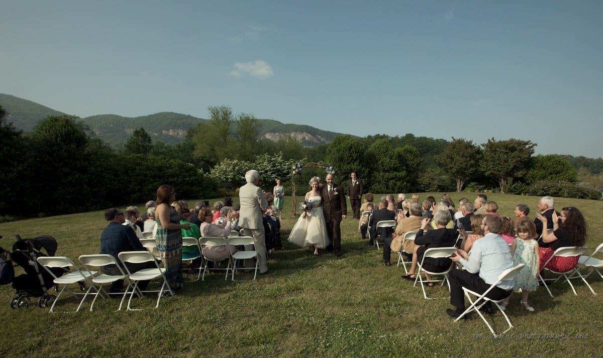 lake lure north carolina wedding photography ~ shannon and paul A bride and groom walk down an aisle outside, surrounded by seated guests on a grassy lawn. The scene is set against a backdrop of green hills under a partly cloudy sky, creating a serene and picturesque wedding setting.