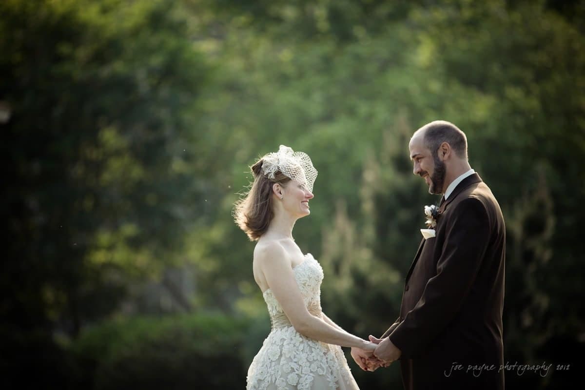 lake lure north carolina wedding photography ~ shannon and paul A bride and groom hold hands and smile at each other outdoors. The bride wears a white dress with a netted fascinator, and the groom is in a dark suit. The background is blurred greenery.