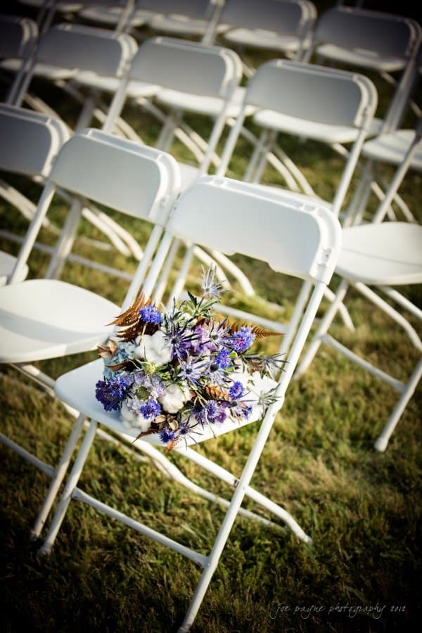 lake lure north carolina wedding photography ~ shannon and paul A bouquet of vibrant purple and white flowers lies on a white folding chair, set against rows of similar chairs on a grassy lawn. The scene suggests an outdoor event, possibly a wedding ceremony.