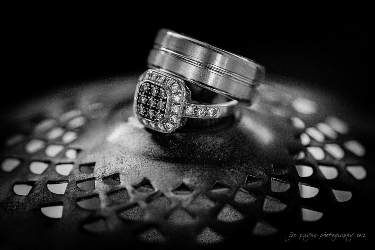 lake lure north carolina wedding photography ~ shannon and paul A black and white photo of a set of wedding rings on an intricate, textured surface. One ring is encrusted with small diamonds, while the other has a plain, smooth design.