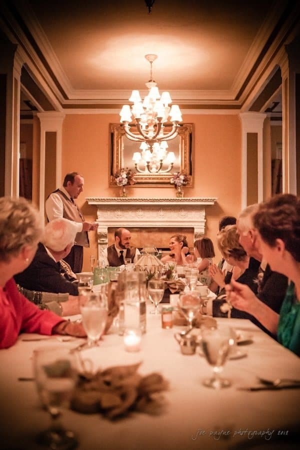 lake lure north carolina wedding photography ~ shannon and paul A group of people sit around a long dining table in a warmly lit room. A man stands at one end of the table, seemingly giving a speech. A chandelier hangs above, and there’s a fireplace with a mirror and flower arrangements behind him.