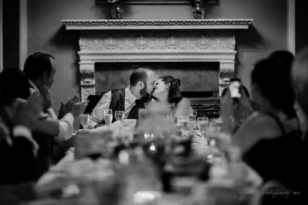 lake lure north carolina wedding photography ~ shannon and paul A black and white photo captures a bride and groom sharing a kiss at their wedding reception. They are seated at a long table with guests applauding around them. A decorated fireplace is in the background.