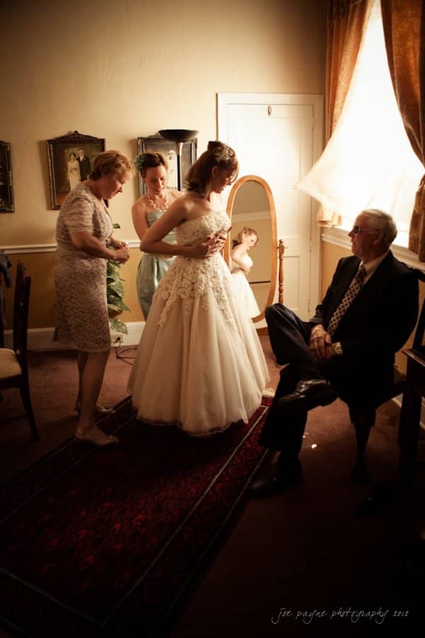 lake lure north carolina wedding photography ~ shannon and paul A bride in a white gown is being assisted by two women in a warmly lit room. A man sits on a chair nearby, watching. A mirror reflects the scene, and there are framed pictures on the wall. The atmosphere is intimate and serene.