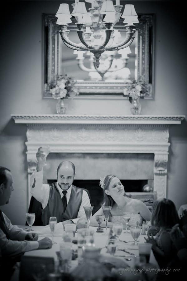 lake lure north carolina wedding photography ~ shannon and paul A black-and-white photo shows a man in formal attire raising his glass at a dining table. A woman in a wedding dress sits beside him, smiling. A chandelier hangs above, and a decorative mirror is in the background.