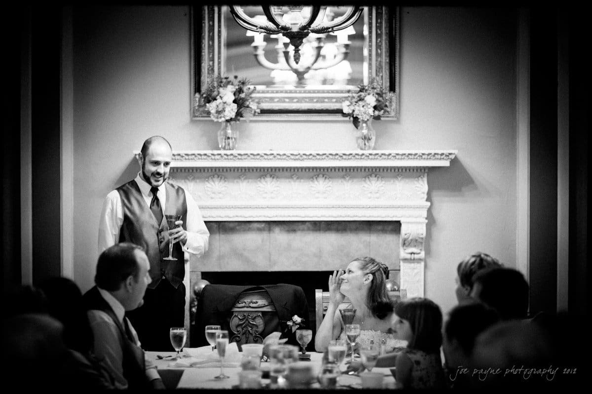 lake lure north carolina wedding photography ~ shannon and paul A man in a vest and tie gives a toast at a formal gathering. He stands near a woman sitting at a table who is smiling up at him. Behind them is a decorative fireplace and a mirror reflecting a chandelier. The mood is celebratory.