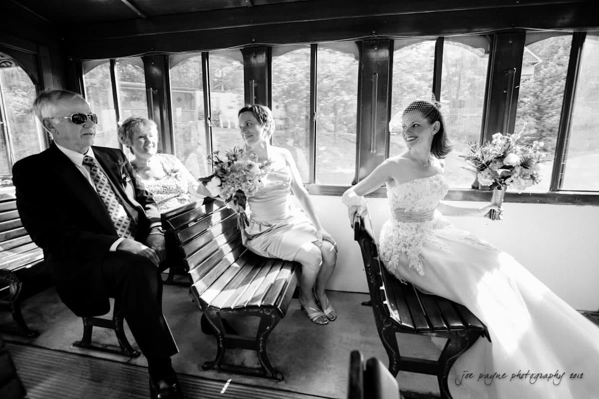 lake lure north carolina wedding photography ~ shannon and paul A bride in a wedding dress sits smiling on a vintage trolley, holding a bouquet. Across from her, three people sit: an older man in a suit, an older woman in floral attire, and a younger woman in a dress holding flowers.