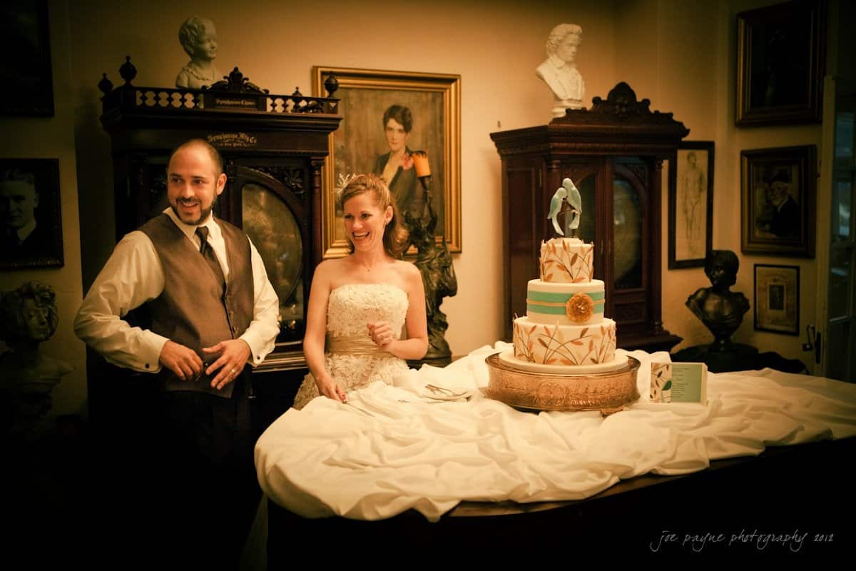lake lure north carolina wedding photography ~ shannon and paul A bride and groom joyfully stand beside a three-tier wedding cake with a decorative topper. They are in a room adorned with classical busts and paintings, draped with a white cloth, creating an elegant and vintage atmosphere.