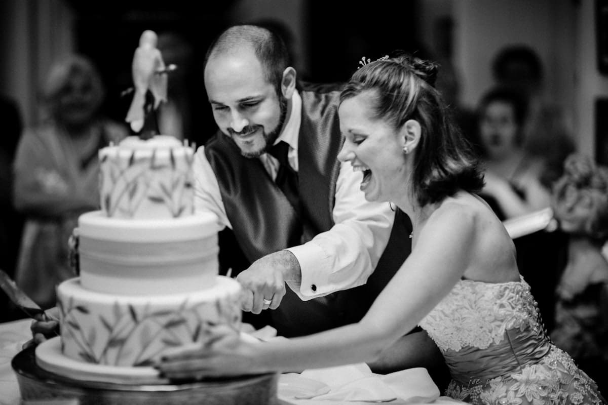 lake lure north carolina wedding photography ~ shannon and paul A smiling couple in wedding attire cuts a decorated tiered cake together. The groom wears a vest and tie, while the bride is in a strapless gown. The scene is celebratory, with onlookers in the background. The image is in black and white.