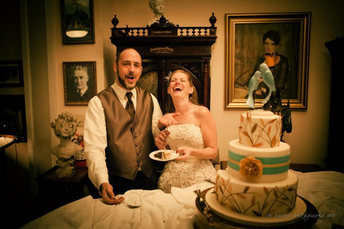 lake lure north carolina wedding photography ~ shannon and paul A couple is celebrating their wedding, standing near a decorated cake. Both are dressed in wedding attire, with the bride laughing joyfully. The room features vintage decor and framed pictures on the walls.