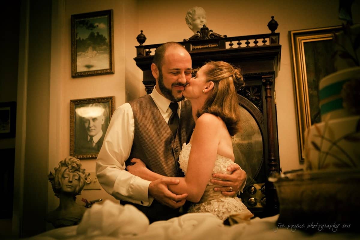lake lure north carolina wedding photography ~ shannon and paul A bride kisses a groom on the cheek in a warmly lit room with vintage decor. They are surrounded by portraits, a statue, and a cake on the right. The bride wears a lace dress, and the groom wears a vest and tie.