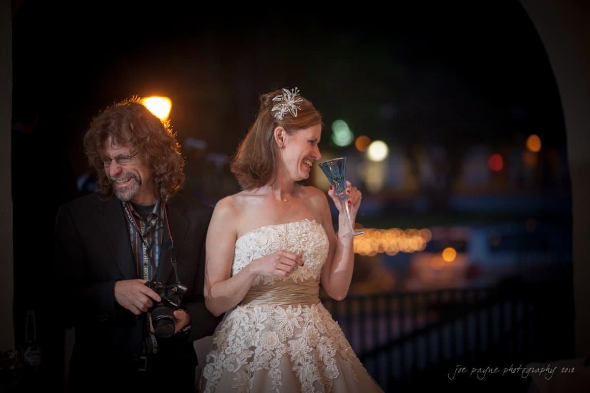 lake lure north carolina wedding photography ~ shannon and paul A bride in a strapless white lace dress smiles while holding a glass, standing next to a man in a black suit who is laughing and holding a camera. They are outdoors, with blurred lights in the background.
