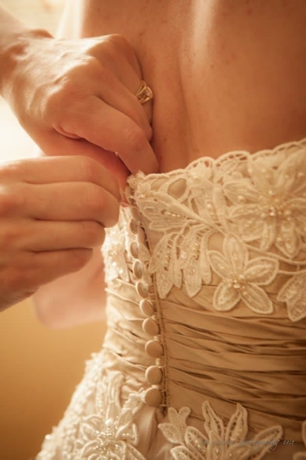 lake lure north carolina wedding photography ~ shannon and paul Close-up of hands fastening the buttons on the back of a lace-embellished wedding dress, creating an elegant pattern of floral details. The scene captures the intricate design and delicate craftsmanship of the bridal gown.