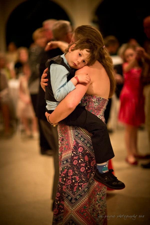 lake lure north carolina wedding photography ~ shannon and paul A woman in a floral dress holds a young boy wearing a blue shirt and vest in her arms. They are surrounded by people dancing in a softly lit room. The boy appears relaxed, resting his head on the womans shoulder.