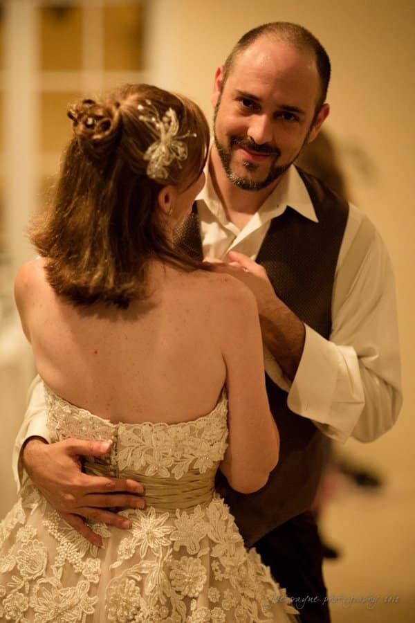 lake lure north carolina wedding photography ~ shannon and paul A couple dances closely at what appears to be a wedding reception. The woman is wearing a strapless lace dress with floral details and a decorative hairpiece. The man is dressed in a vest and white shirt, smiling warmly at her.