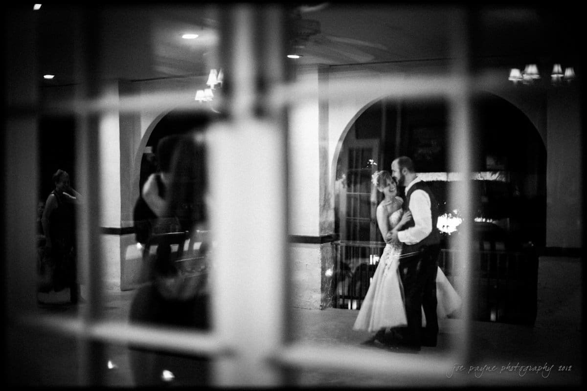 lake lure north carolina wedding photography ~ shannon and paul A couple dances intimately in a dimly lit room, captured through a window with reflective panes. The image has a soft, blurry quality, creating a romantic and nostalgic atmosphere. Other guests appear in the background.