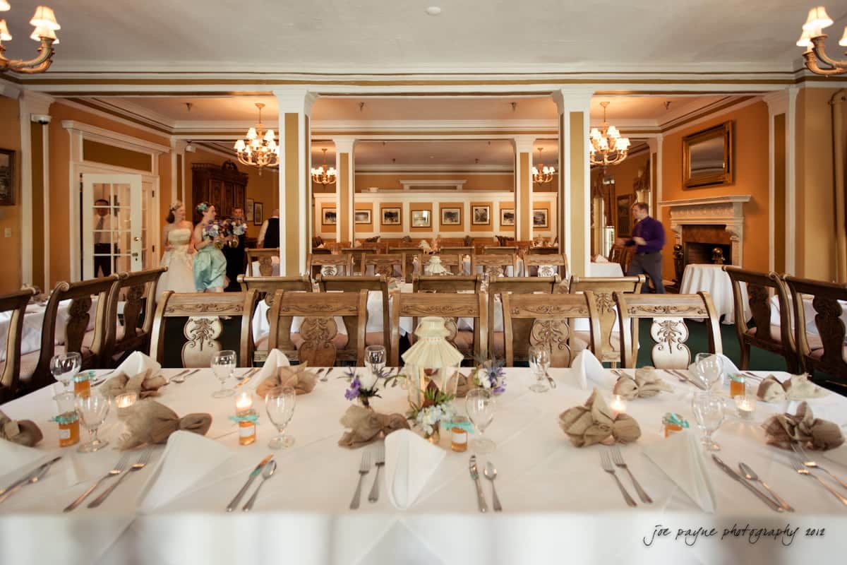lake lure north carolina wedding photography ~ shannon and paul Elegant banquet hall set for an event, featuring long tables with white tablecloths, neatly folded napkins, and decorative centerpieces. Chandeliers illuminate the room, while framed artworks adorn the walls in the background.