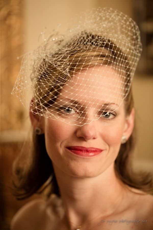lake lure north carolina wedding photography ~ shannon and paul A woman with shoulder-length brown hair is smiling warmly. She is wearing a decorative white netting over her hair and a pearl earring. The background is softly blurred, giving the image a warm, inviting feel.