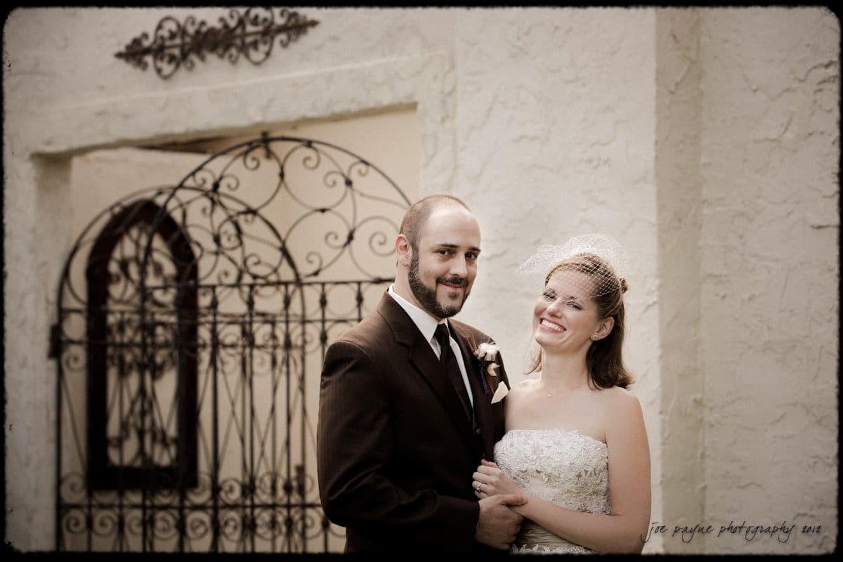 lake lure north carolina wedding photography ~ shannon and paul A bride in a strapless lace gown and birdcage veil smiles while holding hands with a groom in a dark suit and tie. They stand in front of an ornate wrought iron gate against a textured stucco wall.