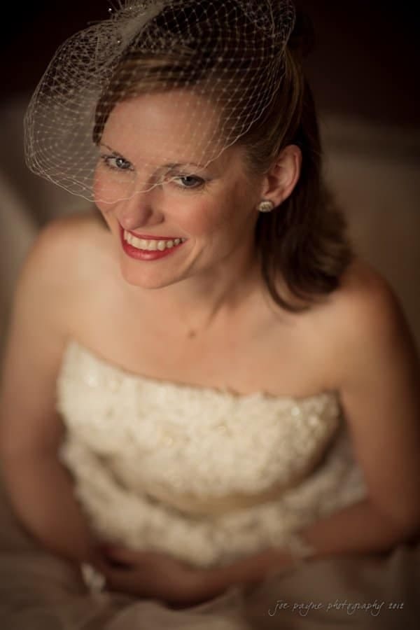 lake lure north carolina wedding photography ~ shannon and paul A woman in a wedding dress is smiling upwards. She has light brown hair and is wearing a birdcage veil on her head. The background is blurred, giving a soft focus to the image.