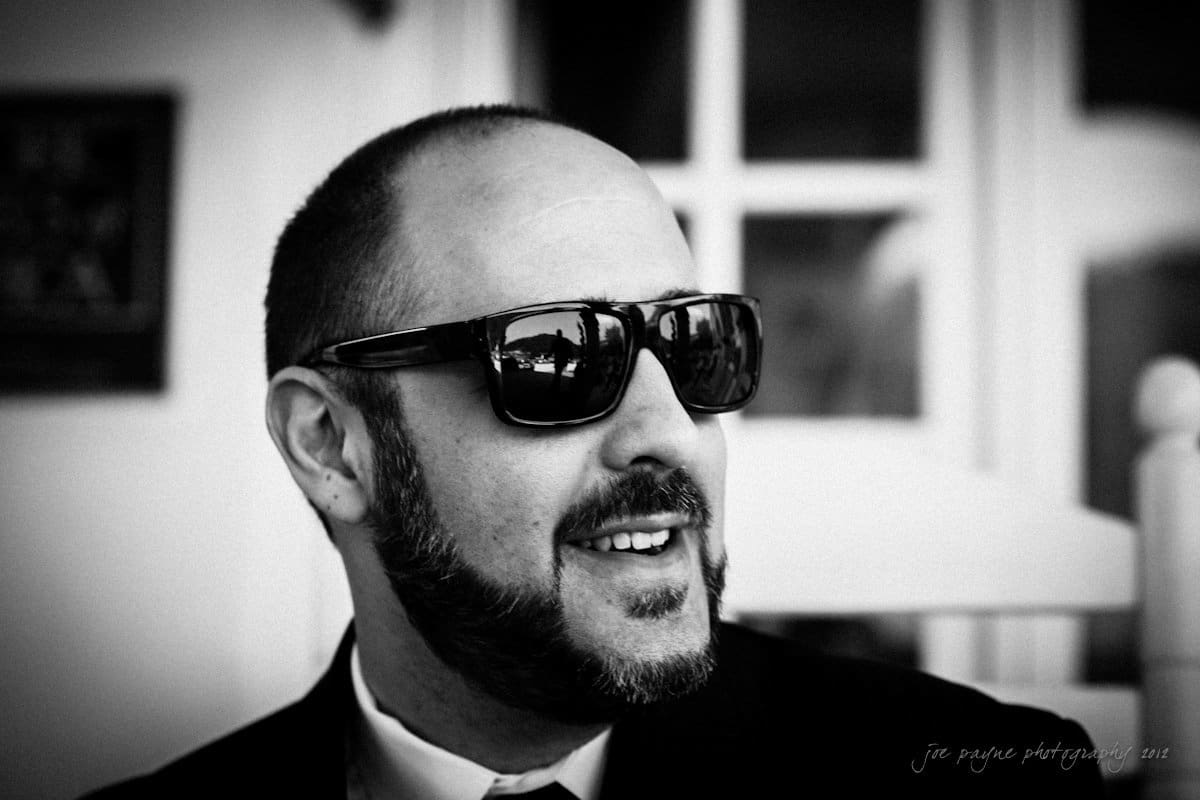 lake lure north carolina wedding photography ~ shannon and paul A black and white photo of a man with a beard, wearing sunglasses and a suit. He is smiling and looking slightly to the side. The background is blurred, focusing attention on the mans face.