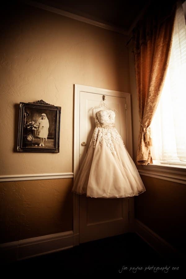 lake lure north carolina wedding photography ~ shannon and paul A vintage wedding dress hangs on a door in a warmly lit room. An old photograph of a child in a dress is framed on the wall. Sunlight streams through a window with long, draped curtains, casting soft shadows.