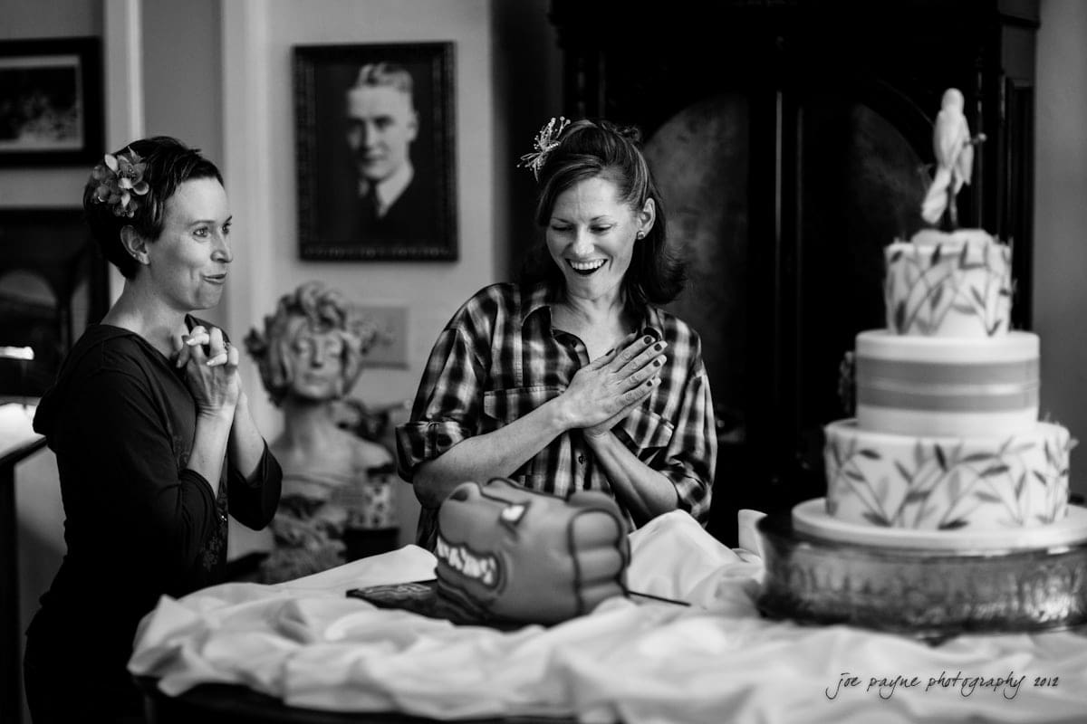 lake lure north carolina wedding photography ~ shannon and paul Two women smile and look at a decorated cake on a table. One is wearing a checkered shirt, and both have flower hair accessories. Theres a portrait on the wall and a bust in the background. The scene is joyful and celebratory.