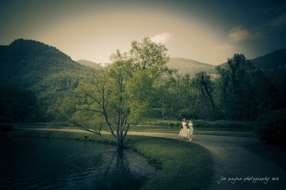 A serene landscape with a small lake surrounded by lush greenery and rolling hills under a cloudy sky. Two people in formal attire walk together along a curved path beside the water. The scene has a dreamy, vintage aesthetic.