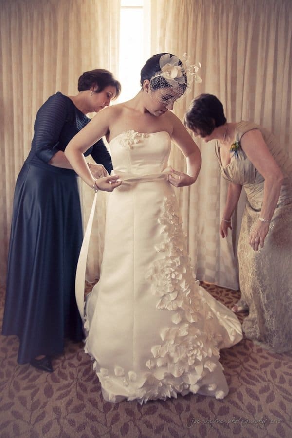 A bride stands adjusting her white strapless wedding dress adorned with floral appliques and a birdcage veil. Two women, both elegantly dressed, assist her with the gown in a warmly lit room with patterned carpet and draped curtains.