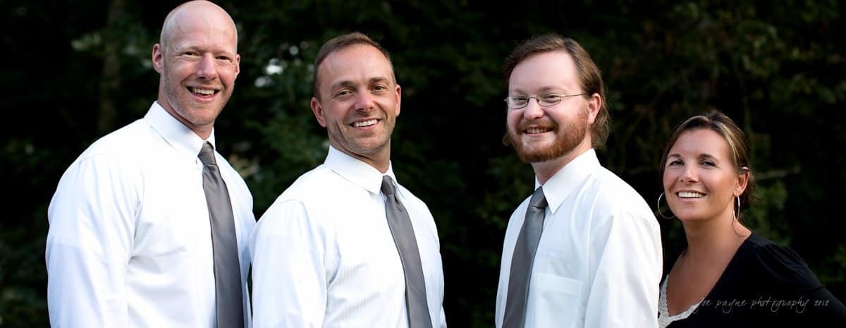 A group of four people, three men and one woman, stand outdoors smiling. They are wearing white shirts and gray ties, and the woman is wearing a black top. The background is filled with dark green foliage.