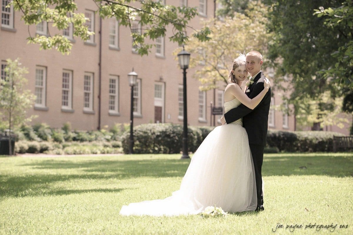 carolina inn wedding photography ~ danielle and thomas A bride and groom embrace on a sunlit lawn in front of a large building. The bride wears a white gown and the groom a black suit. Trees and a lamppost are in the background, creating a serene and romantic setting.