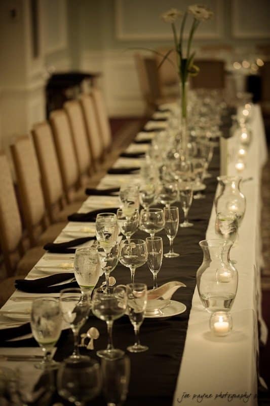 carolina inn wedding photography ~ danielle and thomas An elegant, elongated dining table is set with white plates, glassware, and black table runners. The table features clear vases and candles, with simple floral arrangements. Beige chairs line both sides in a softly lit room.