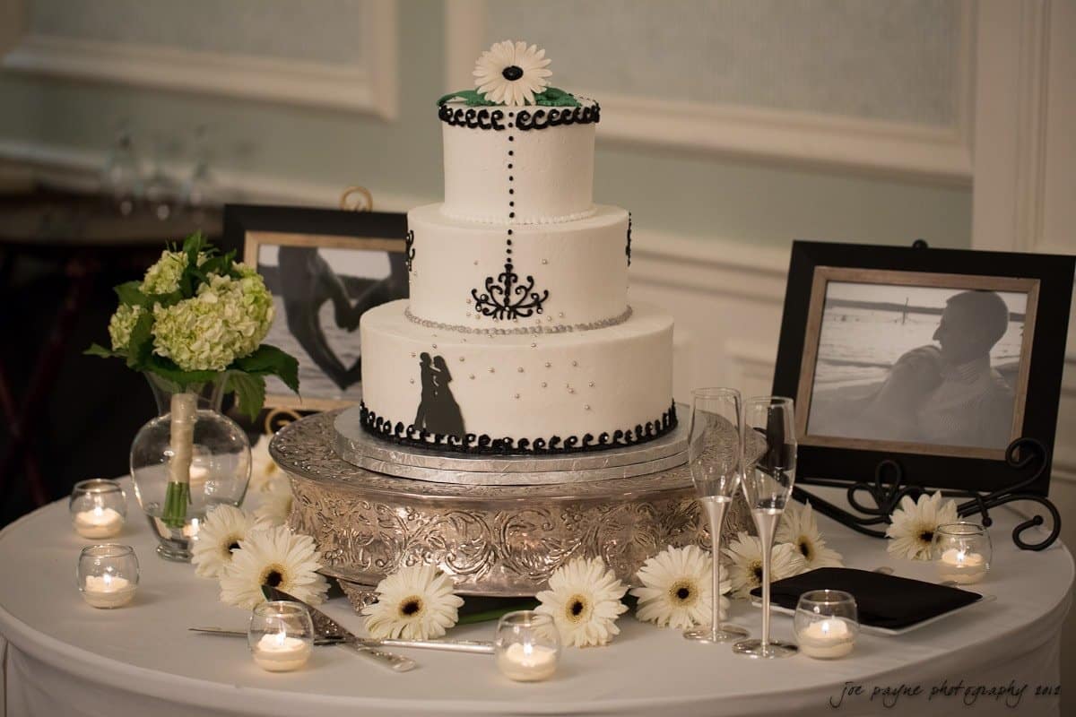 carolina inn wedding photography ~ danielle and thomas A three-tiered white wedding cake with black accents and a bride-groom silhouette topper, surrounded by white flowers and candles. Framed photos and a vase with green hydrangeas are also on the decorated table.
