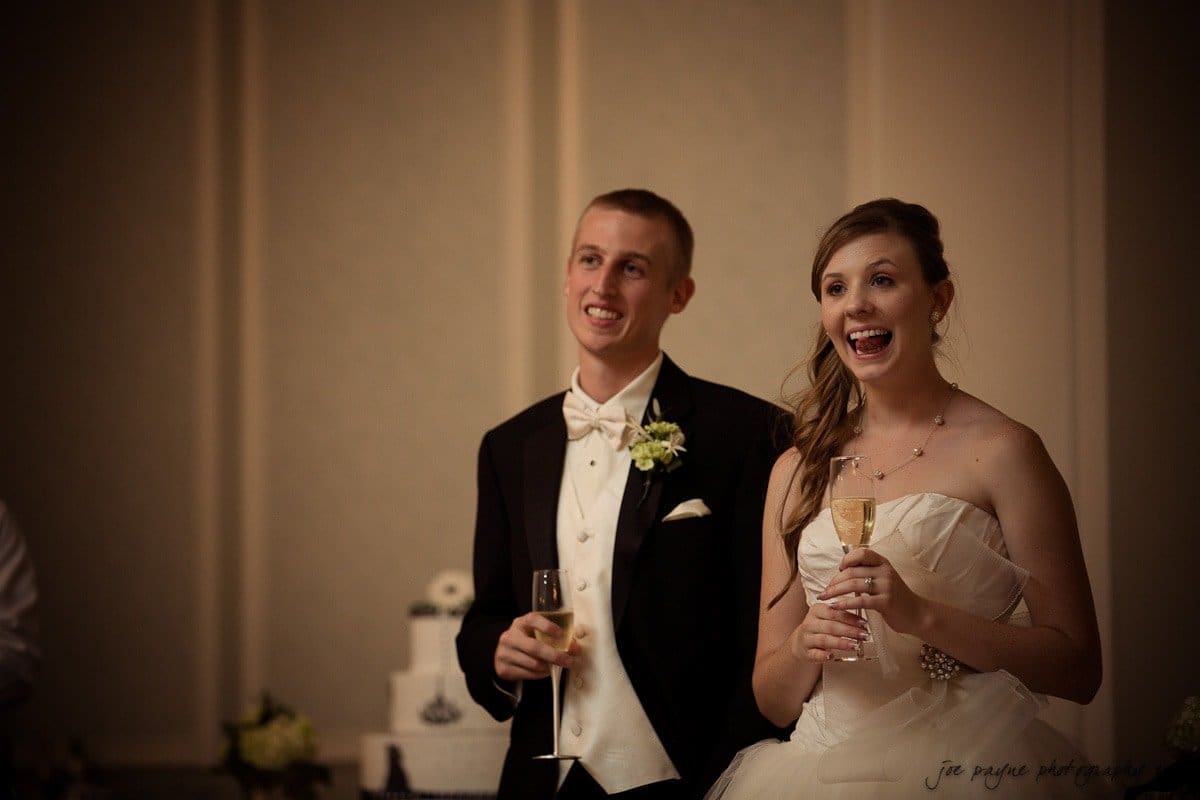 carolina inn wedding photography ~ danielle and thomas A bride and groom are standing together in formal attire at a wedding reception. Both are smiling and holding champagne glasses. A wedding cake is visible in the background.