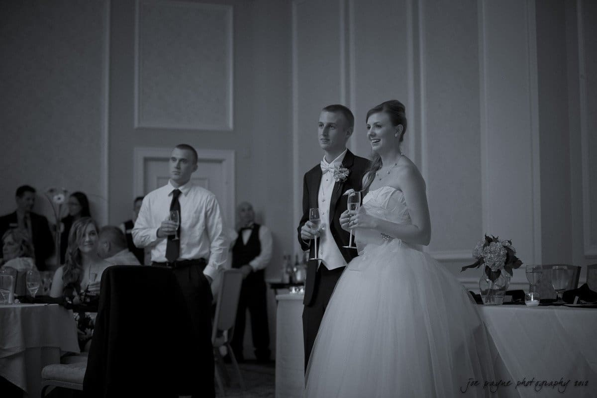 carolina inn wedding photography ~ danielle and thomas A bride and groom stand in a formal setting, both holding glasses. The bride is in a wedding dress and the groom in a tuxedo. Guests are seated around them, some with drinks in hand. The room has elegant decor with chandeliers and paneling.
